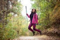 Hiker girl jumping on a trail in the mountains. Backpacker with hiking poles and pink jacket in a forest. Happy lifestyle outdoors