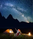 Hiker man showing his lady stars and Milky way in the night sky. Couple sitting near the lighting tent and campfire.