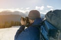 Hiker man tourist photographer in warm clothing with backpack and camera taking picture of snowy valley and woody mountain peaks Royalty Free Stock Photo