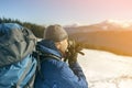 Hiker man tourist photographer in warm clothing with backpack and camera taking picture of snowy valley and woody mountain peaks Royalty Free Stock Photo