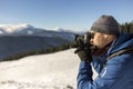 Hiker man tourist photographer in warm clothing with backpack and camera taking picture of snowy valley and woody mountain peaks Royalty Free Stock Photo