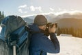 Hiker man tourist photographer in warm clothing with backpack and camera taking picture of snowy valley and woody mountain peaks Royalty Free Stock Photo