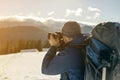 Hiker man tourist photographer in warm clothing with backpack and camera taking picture of snowy valley and woody mountain peaks Royalty Free Stock Photo