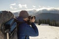 Hiker man tourist photographer in warm clothing with backpack and camera taking picture of snowy valley and woody mountain peaks Royalty Free Stock Photo