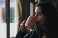 Hiker portrait with pink cup in the hands looking through the window. Woman drinking hot tea from a mug in a mountain shelter