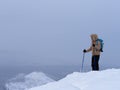 Hiker in a jacket with a fur hood, backpack and nordic walking sticks stands in the snow on the mountains in winter