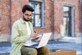 Hindu student in headphones sitting outside on a bench, holding a laptop on his lap, writing in a notebook, studying Royalty Free Stock Photo