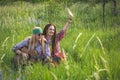 Hippie-style guy and a girl with a guitar are sitting in the grass on a forest glade on a sunny day and calling friends Royalty Free Stock Photo