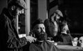 Hipster with beard covered with cape trimming by professional barber in stylish barbershop. Barber busy with grooming Royalty Free Stock Photo