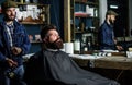 Hipster bearded client got hairstyle. Barber with hairdryer blows off hair out of cape. Barber with hairdryer works on Royalty Free Stock Photo