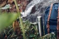 Hipster Blue Backpack, Trekking Poles And Thermos Closeup. View From Front Tourist Traveler Bag On Waterfall Background