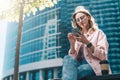 Hipster businesswoman in glasses sits outdoors and uses smartphone. Nearby is cup of coffee. In background is skyscraper Royalty Free Stock Photo