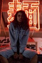 Hipster cool African gen z lady looking at camera sit under red neon night sign.