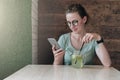 Hipster girl in glasses sits in cafe at table, drinks beverage and uses smartphone. Businesswoman working, blogging Royalty Free Stock Photo