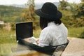 Hipster girl with laptop sitting on wooden porch with beautiful view on woods and mountains. Stylish Young woman in hat using