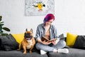 Hipster girl sitting on sofa near small french bulldog and reading book Royalty Free Stock Photo