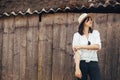 Hipster girl in white casual outfit posing on background of old wooden cabin in mountains. Stylish woman in hat standing near old