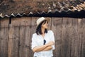Hipster girl in white casual outfit posing on background of old wooden cabin in mountains. Stylish woman in hat standing near old