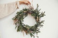 Hipster girl in white sweater holding rural christmas wreath with fir branches, berries, pine cones and herbs  on white wall in Royalty Free Stock Photo