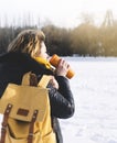 Hipster girl with yellow backpack drinking hot tea or coffee on background of winter forest in the snow mountains. Hiking tourist