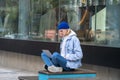 Hipster guy in trendy outwear sitting in street, working freelance online, messaging in internet