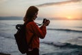 Hipster hiker tourist with backpack making photo of seascape sunset on camera on background sea, photographer enjoying ocean horiz