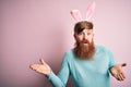 Hipster Irish man with beard wearing easter rabbit ears over isolated pink background clueless and confused expression with arms