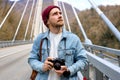 Hipster male in denim jacketwalking on bridge in countryside using camera