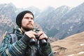 A hipster man with a beard in a hat, a jacket, and a backpack in the mountains holds binoculars, adventure, tourism