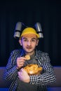 Hipster man with beer helmet on the head eating chips while watching TV At home Royalty Free Stock Photo