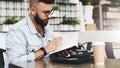 Hipster man in glasses is sitting in cafe, reading notes in notebook. On table is laptop,cup of coffee, instant camera. Royalty Free Stock Photo