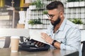 Hipster man in glasses is sitting in cafe, reading notes in notebook. On table is laptop,cup of coffee, instant camera. Royalty Free Stock Photo