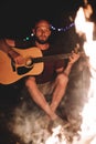 Hipster man playing on acoustic guitar and singing song at big bonfire at camp, chilling with friends in the forest. Group of Royalty Free Stock Photo