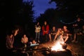 Hipster man playing on acoustic guitar and singing song with friends travelers at big bonfire at night camp in the forest. Group