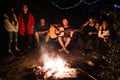 Hipster man playing on acoustic guitar and singing song with friends travelers at big bonfire at night camp in the forest. Group