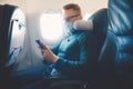 Hipster man in protection mask is watching video on mobile phone, reading online book traveling on airplane Royalty Free Stock Photo