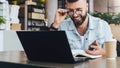 Hipster man sits in cafe, uses smartphone, works on laptop. Businessman reads information message on computer screen.