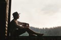 Hipster man sitting on porch of wooden house looking at mountai