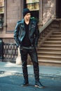 Hipster man wearing black style leather outfit with hat, pants, jacket and shoes standing on city street. Royalty Free Stock Photo