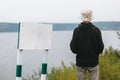 Hipster teenager in bucket hat standing on top of rock mountain with beautiful view on river near empty sign board. Wanderlust and Royalty Free Stock Photo