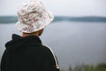 Hipster teenager in bucket hat standing on top of rock mountain and looking on river, view from back. Atmospheric calm moment.