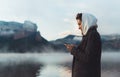 Hipster tourist using smartphone planning trip on foggy lake, mist in mountain nature, traveler girl hold mobile phone, enjoy wifi