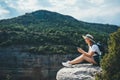 Hipster traveler in hat and backpack ralaxing in nature and using internet on smartphone, tourist girl chatting on cellphone