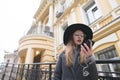 A hipsters girl with a hat and glasses looks at the phone on the background of a beautiful house. Royalty Free Stock Photo