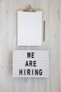 `We are hiring` words on a modern board, clipboard with blank sheet of paper on a white wooden background, top view. Overhead,