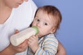 Hirizontal shot of liitle charming baby drinking milk of bottle, being in mother`s arms, feels hunger, looking aside, wearing Royalty Free Stock Photo