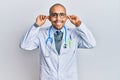 Hispanic adult man wearing doctor uniform and stethoscope smiling pulling ears with fingers, funny gesture