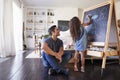 Hispanic dad sitting on the floor in sitting room watching his young daughter drawing on blackboard