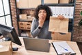 Hispanic doctor woman with curly hair working at small business ecommerce holding paper bag covering mouth with hand, shocked and Royalty Free Stock Photo