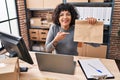 Hispanic doctor woman with curly hair working at small business ecommerce holding paper bag smiling happy pointing with hand and Royalty Free Stock Photo